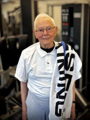 Ein älterer Mann mit Brille und weißem Hemd lächelt mit einem Handtuch über der Schulter in einem hellen Kraftraum mit Trainingsmaschinen.