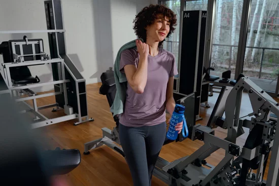 Femme souriante aux cheveux bouclés tenant une gourde bleue dans la salle de musculation, à côté d'appareils d'entraînement gris.