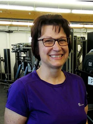 Porträtaufnahme einer lächelnden Frau mit Brille in einem Fitnessstudio, im Hintergrund sind medizinische Krafttrainingsgeräte erkennbar.