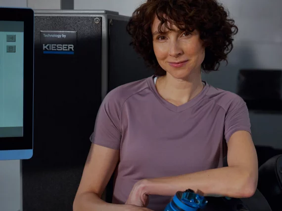 Portrait d'une femme souriante aux cheveux bruns bouclés devant un appareil de musculation gris portant le logo de l'entreprise.
