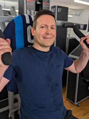 Lächelnder Mann im blauen Oberteil sitzt an einer Trainingsmaschine in einem hellen Kraftraum.