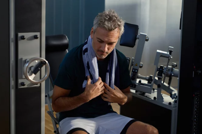 Trainierende Person sitzt nach einer Trainingseinheit auf einem Krafttrainingsgerät in einem Fitnessstudio und hat ein Handtuch über die Schultern gelegt.