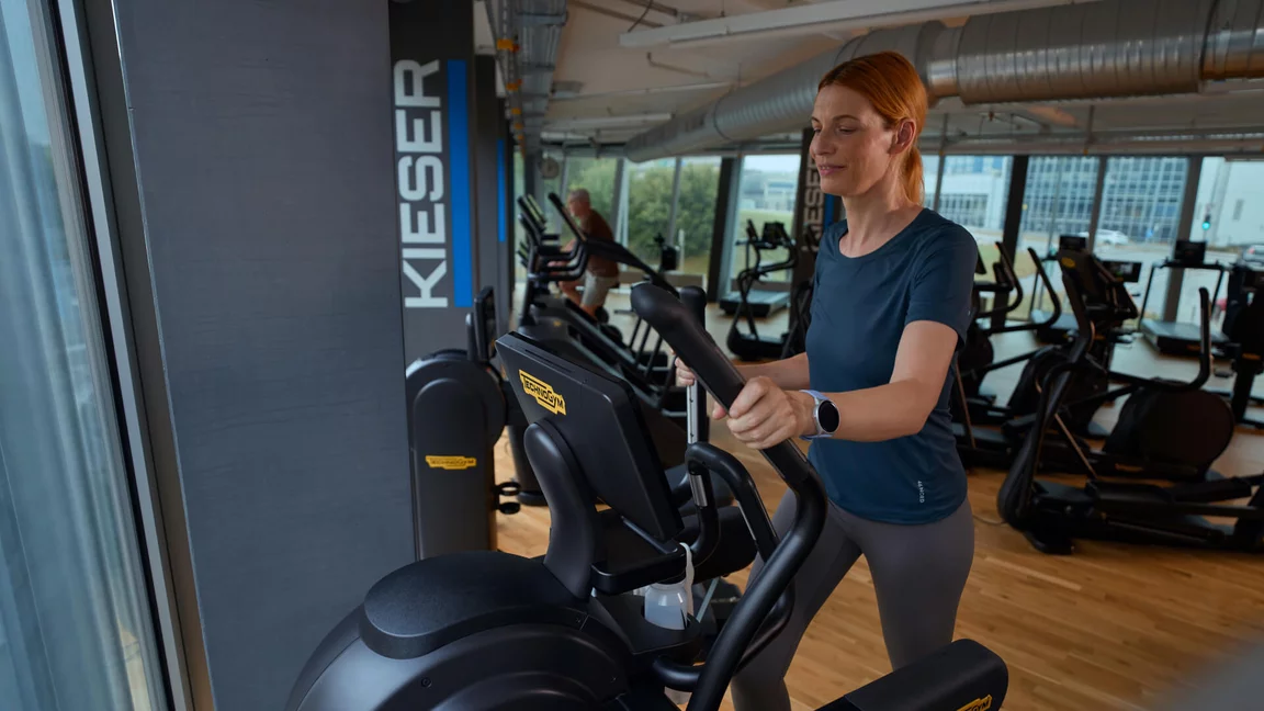 Eine Frau beim Herz-Kreislauf-Training auf einem Crosstrainer in einem modernen, hellen Kieser Studio mit Blick nach draußen.