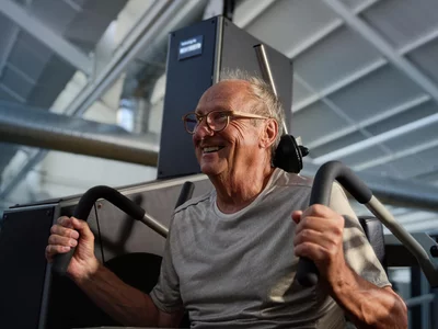 Ein älterer Mann mit Brille lacht bei einer kräftigenden Rückenübung an einer Trainingsmaschine zur Vorbeugung von Rückenschmerzen.