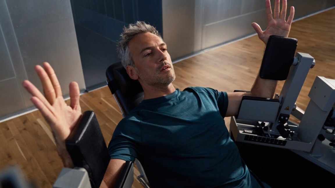 A person is performing an exercise on a strength training machine. Their arms are raised at their sides whilst they work out on a training seat in a Kieser Studio.