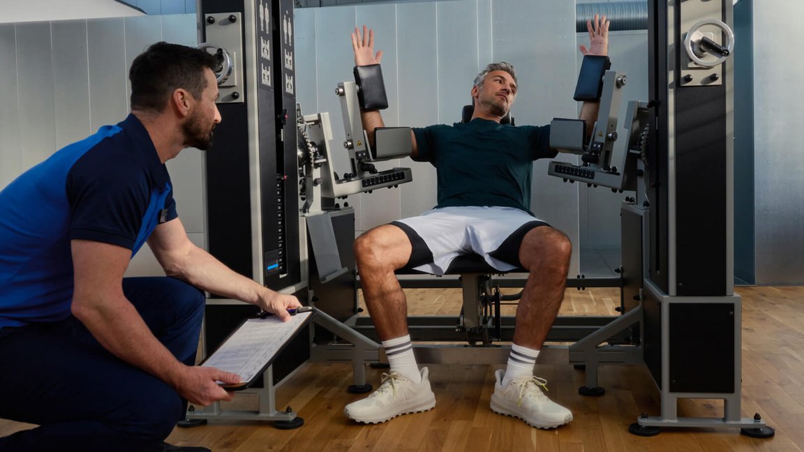 Ein Trainierender an einer spezialisierten E4/5-Maschine erhält professionelle Begleitung durch einen Kieser Instruktor für korrektes Krafttraining.