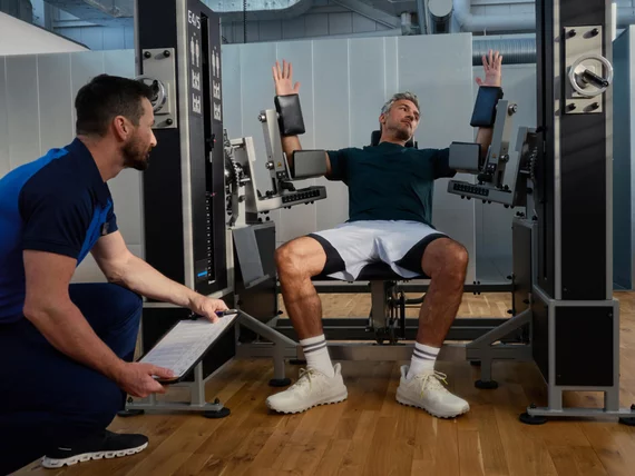 Ein Trainierender an einer spezialisierten E4/5-Maschine erhält professionelle Begleitung durch einen Kieser Instruktor für korrektes Krafttraining.