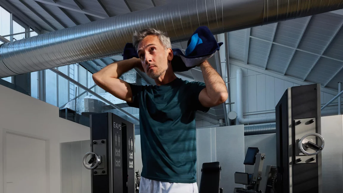 Sportlicher Mann mit graumeliertem Haar im dunkelgrünen T-Shirt trocknet sich mit einem blauem Handtuch ab, im Hintergrund spezialisierte Trainingsmaschinen und eine helle Industriehalle.