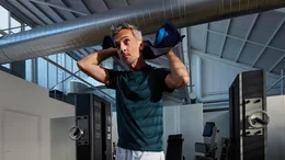 Sportlicher Mann mit graumeliertem Haar im dunkelgrünen T-Shirt trocknet sich mit einem blauem Handtuch ab, im Hintergrund spezialisierte Trainingsmaschinen und eine helle Industriehalle.